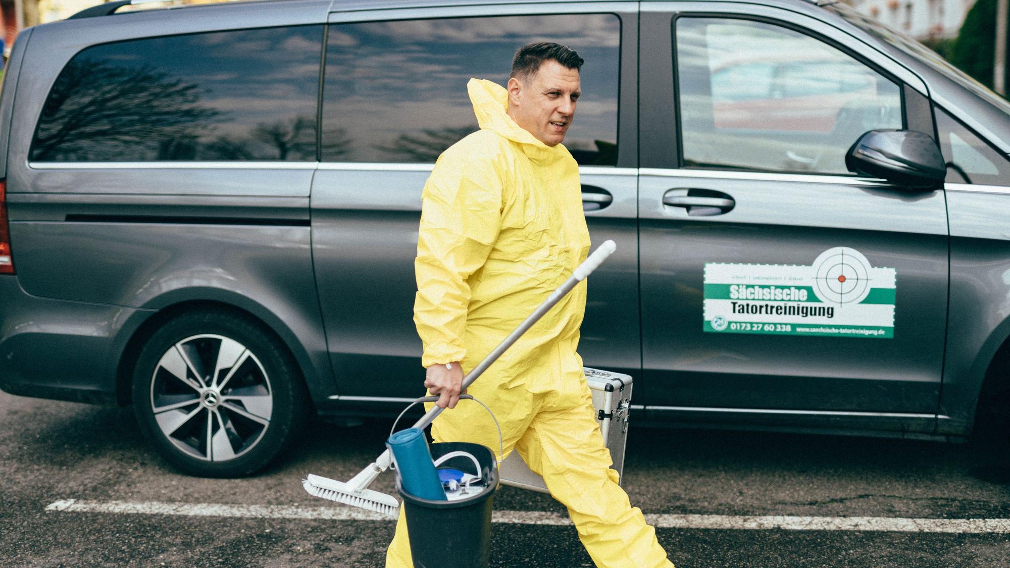 Zu sehen ist Thomas Kundt, der einen gelben Schutzanzug trägt und Putzutensilien mit sich hat. Im Hintergrund ist ein großes Auto auf dem „Sächsische Tatortreinigung“ steht.