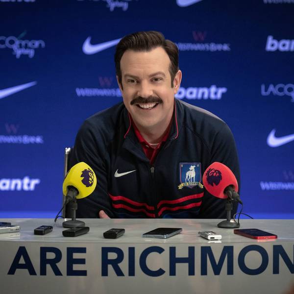 Ted Lasso (Jason Sudeikis) sitzt in der ersten Folge der dritten Staffel der gleichnamigen Serie in einer Pressekonferenz. Er trägt einen dunkelblauen Trainingsanzug.
