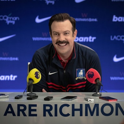 Ted Lasso (Jason Sudeikis) sitzt in der ersten Folge der dritten Staffel der gleichnamigen Serie in einer Pressekonferenz. Er trägt einen dunkelblauen Trainingsanzug.