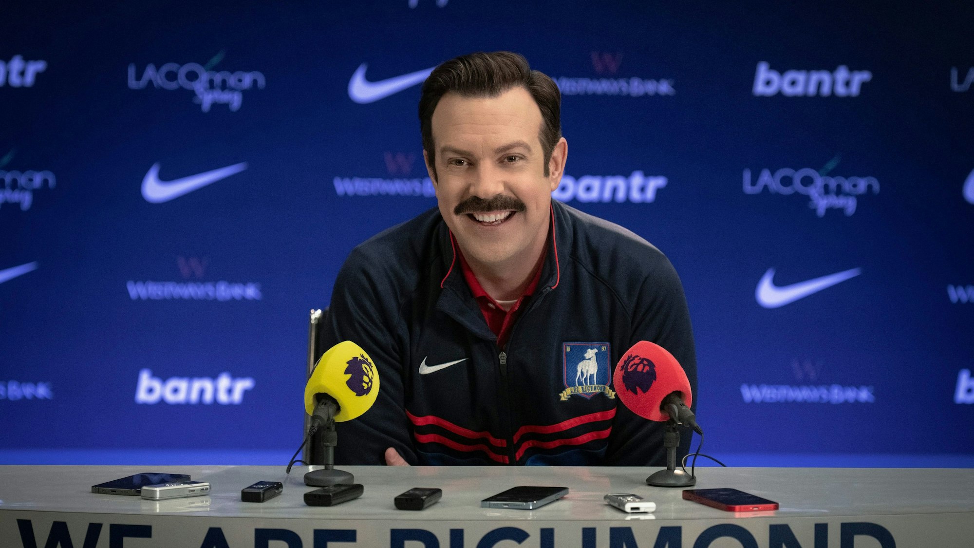 Ted Lasso (Jason Sudeikis) sitzt in der ersten Folge der dritten Staffel der gleichnamigen Serie in einer Pressekonferenz. Er trägt einen dunkelblauen Trainingsanzug.
