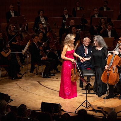 Anne-Sophie Mutter trägt ein magentafarbenes Kleid und gibt Martha Argerich die Hand. Daneben sitzt Mischa Maisky mit seinem Cello.