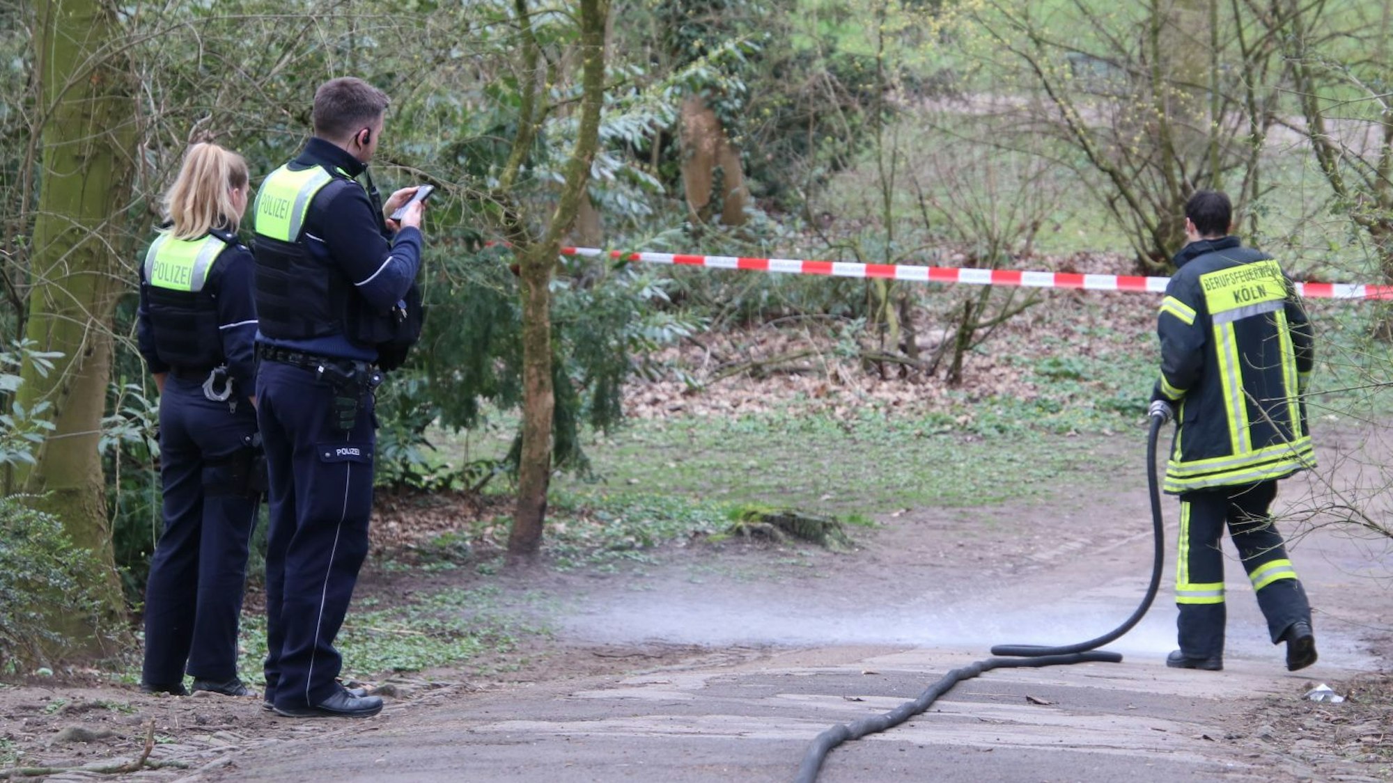 Zwei Polizisten und ein Feuerwehrmann stehen vor einem Absperrband im Weidenpescher Park.