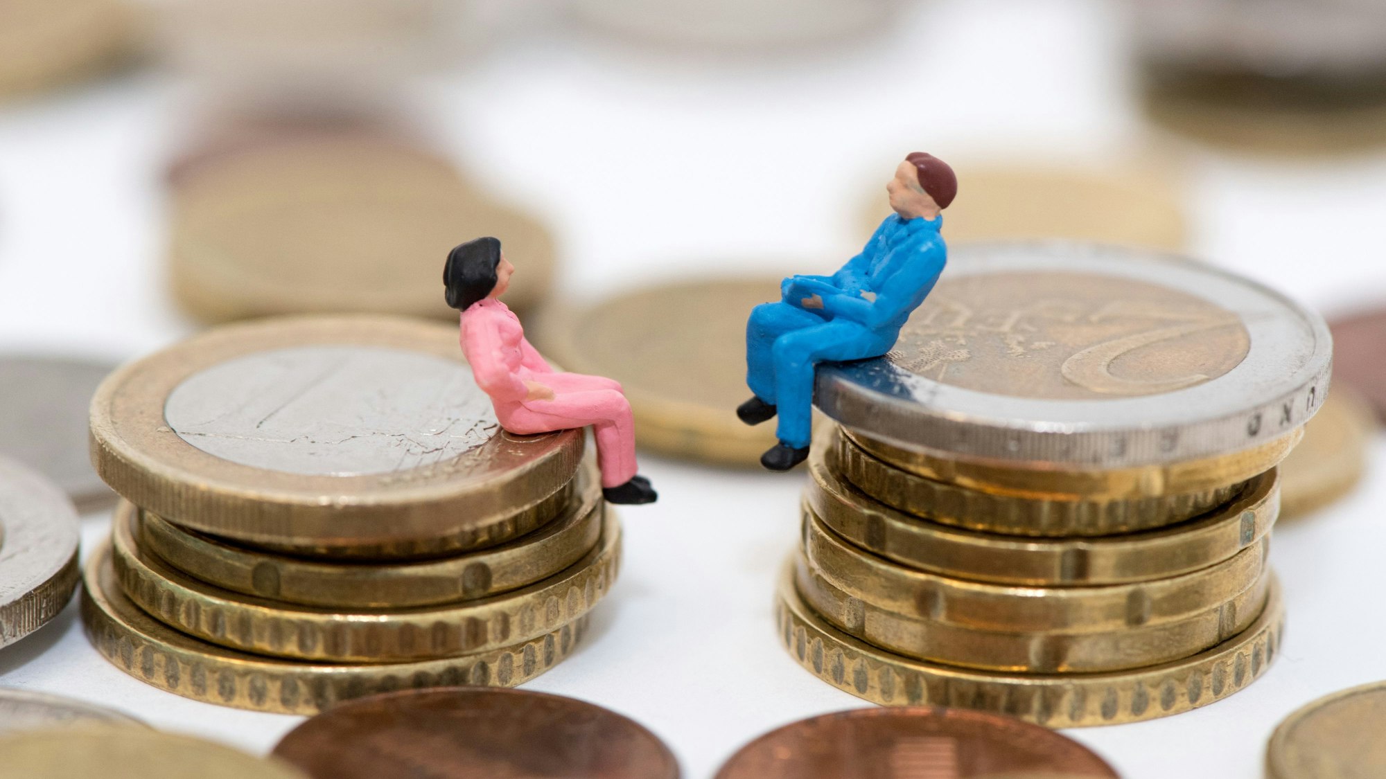 Zwei kleine Figuren, ein Mann und eine Frau, sitzen auf einem Stapel Geldmünzen
