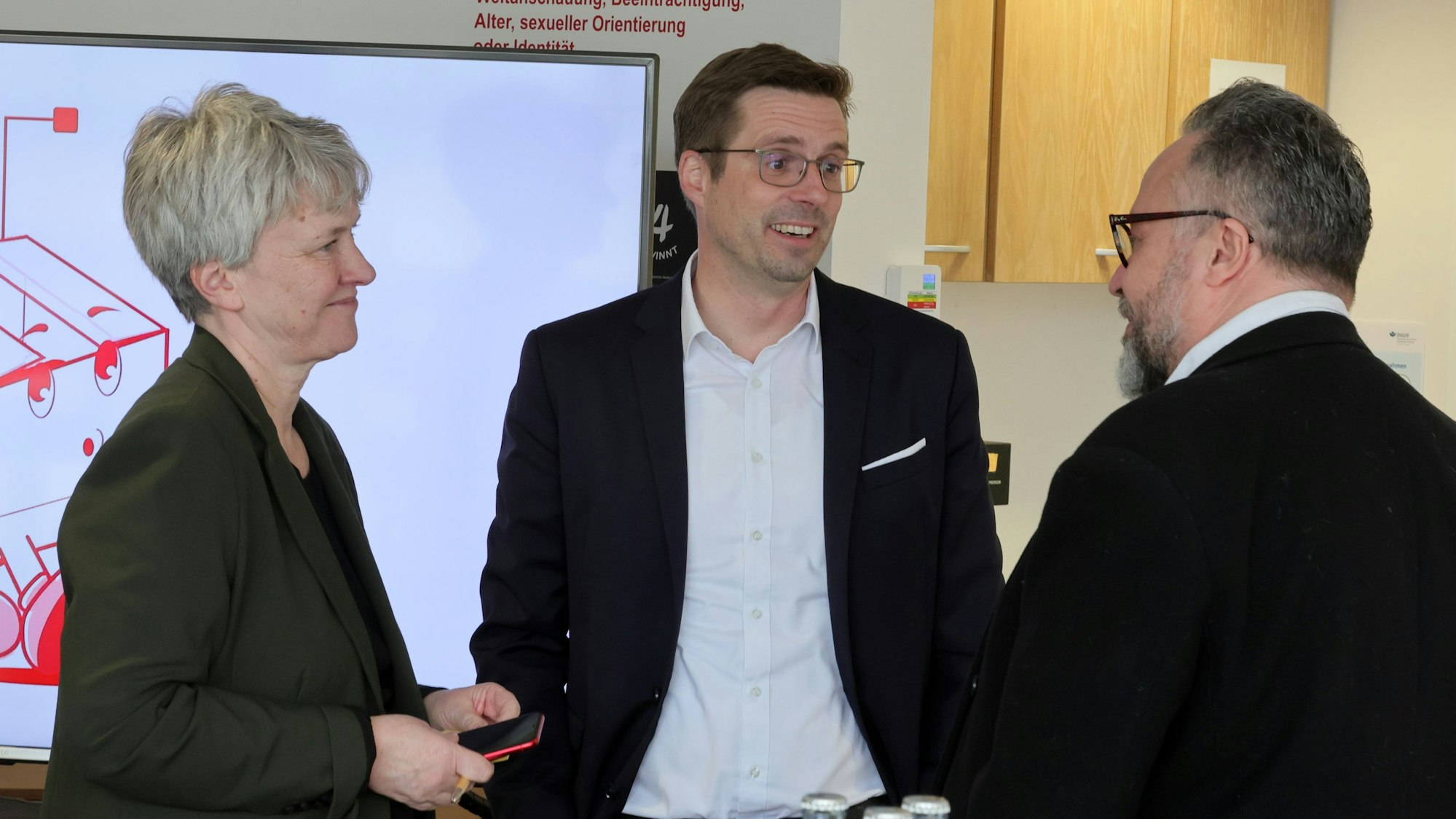 Sabine Möwes, Andree Haack und Dirk Blauhut stehen vor einem Fernseher mit einer Präsentation des Projekts.
