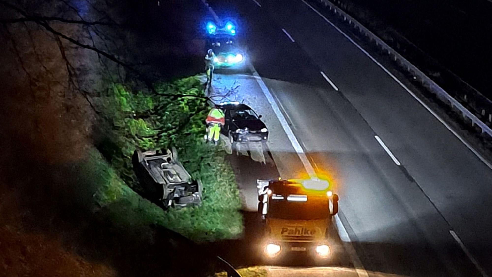 Einsatzfahrzeuge mit Blaulicht und Gelblicht stehen auf dem Seitenstreifen einer Autobahn. Ein Unfallwagen liegt auf dem Dach im Straßengraben. Ein weiteres Unfallauto steh ebenfalls auf dem Seitenstreifen.
