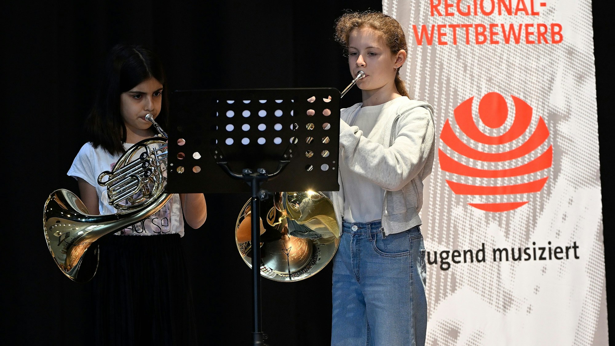 Zwei junge Hornistinnen musizieren vor einem Banner des Regionalwettbewerbs „Jugend musiziert“.