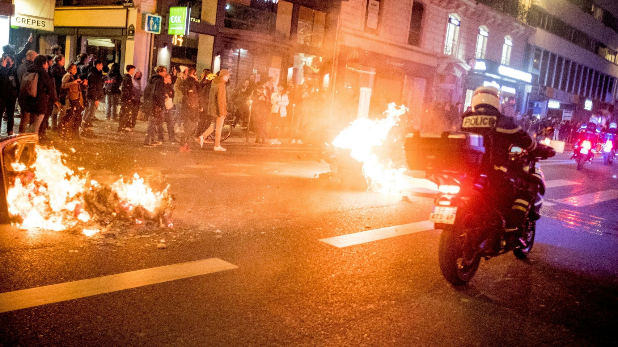 Ein Polizist fährt auf einem Motorrad an brennenden Mülltonnen in der französischen Hauptstadt Paris vorbei. In der Nacht zu Dienstag sind aufgrund der verabschiedeten Rentenreform Hunderte Menschen auf die Straßen gegangen.