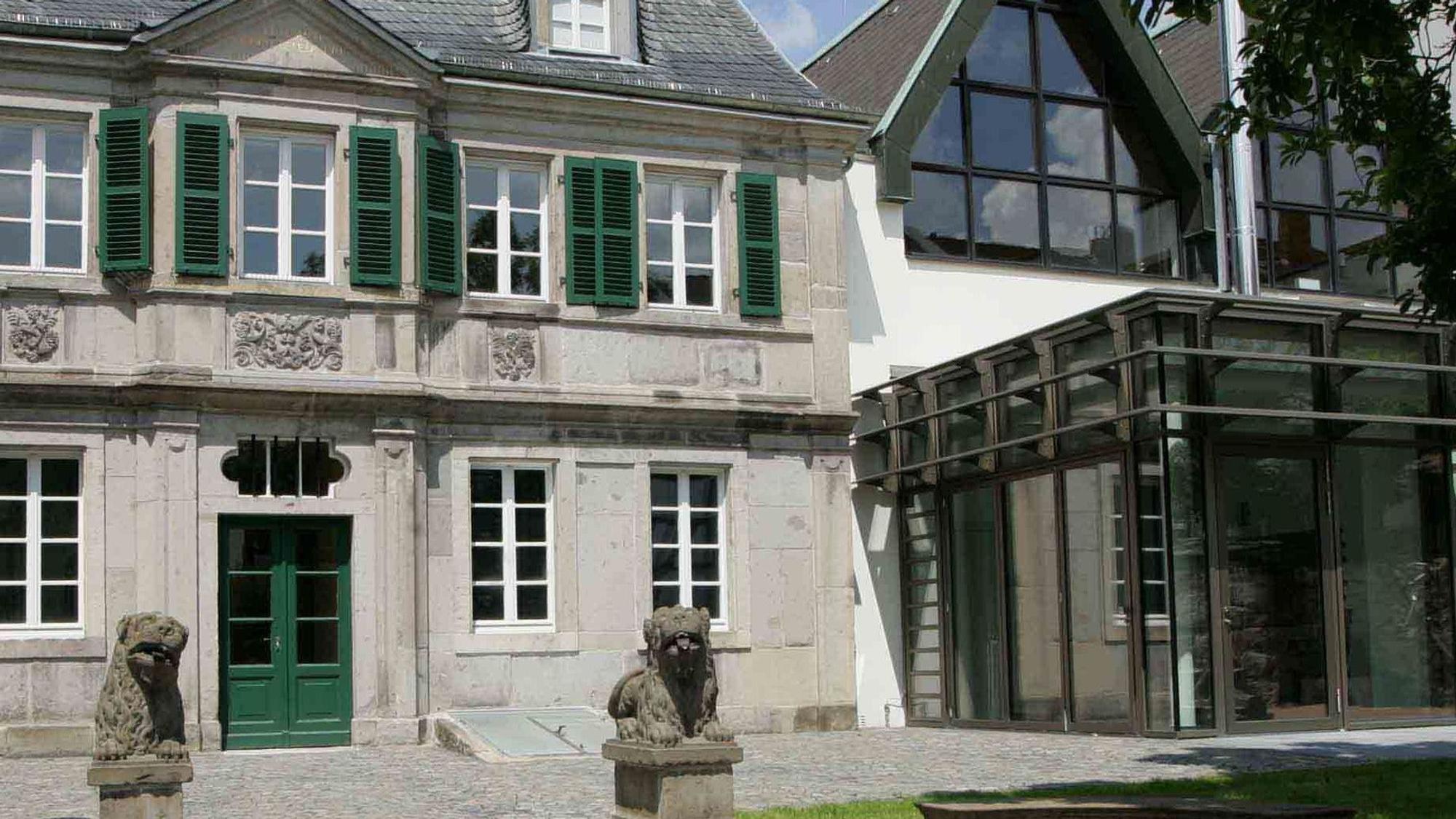 Zwei Steinlöwen sitzten auf Säulen vor einem Barockgebäude, dessen Tür und Fensterläden grün angestrichen sind. Rechs daneben befindet sich ein moderner Glasbau, um den das Museum erweitert worden ist.