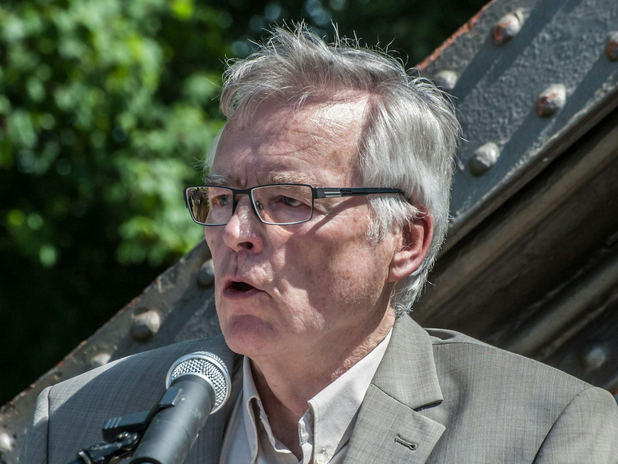 Ein Mann mit eckiger Metallbrille und grauem Anzug spricht in ein Mikrofon.
