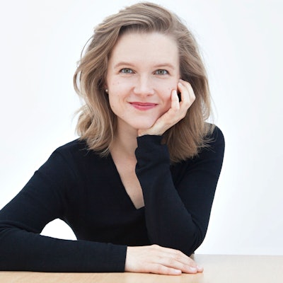 Die Dirigentin lächelt in die Kamera. Sie sitzt und stützt ihr Kinn mit ihrer Hand, Ellebogen am Tisch angelehnt.