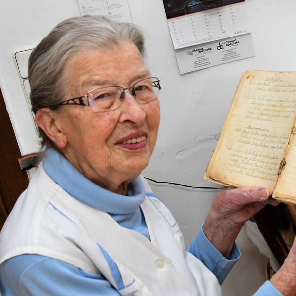 Oma Agnes sitzt an ihrem Küchentisch und zeigt ihr Rezeptbuch. Auf der Tischplatte liegen ausgerollter Teig und ein Nudelholz.