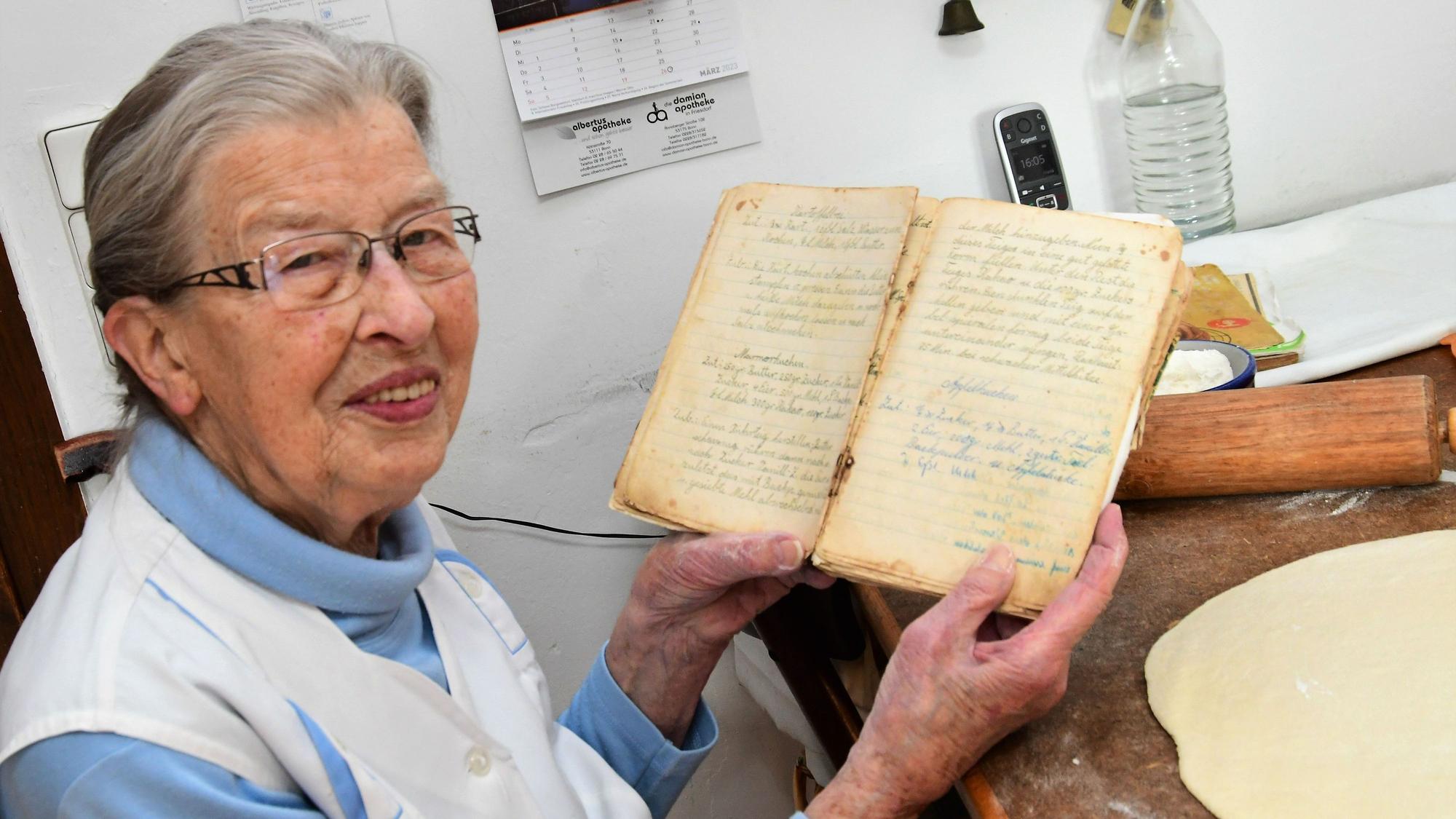 Oma Agnes sitzt an ihrem Küchentisch und zeigt ihr Rezeptbuch. Auf der Tischplatte liegen ausgerollter Teig und ein Nudelholz.