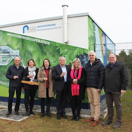 SIeben Personen stehen vor einem Gebäude und halten Plastikbecher mit Wasser in der Hand. Eine bemalte Hauswand im Hintergrund weist auf die Wasserwerkskooperation hin.