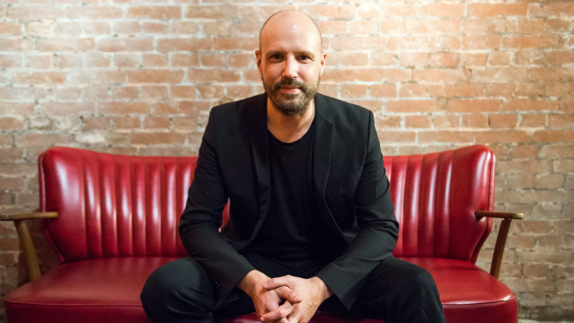 Christopher von Deylen sitzt im schwarzen Anzug auf einer roten Bank vor einer Backsteinmauer.