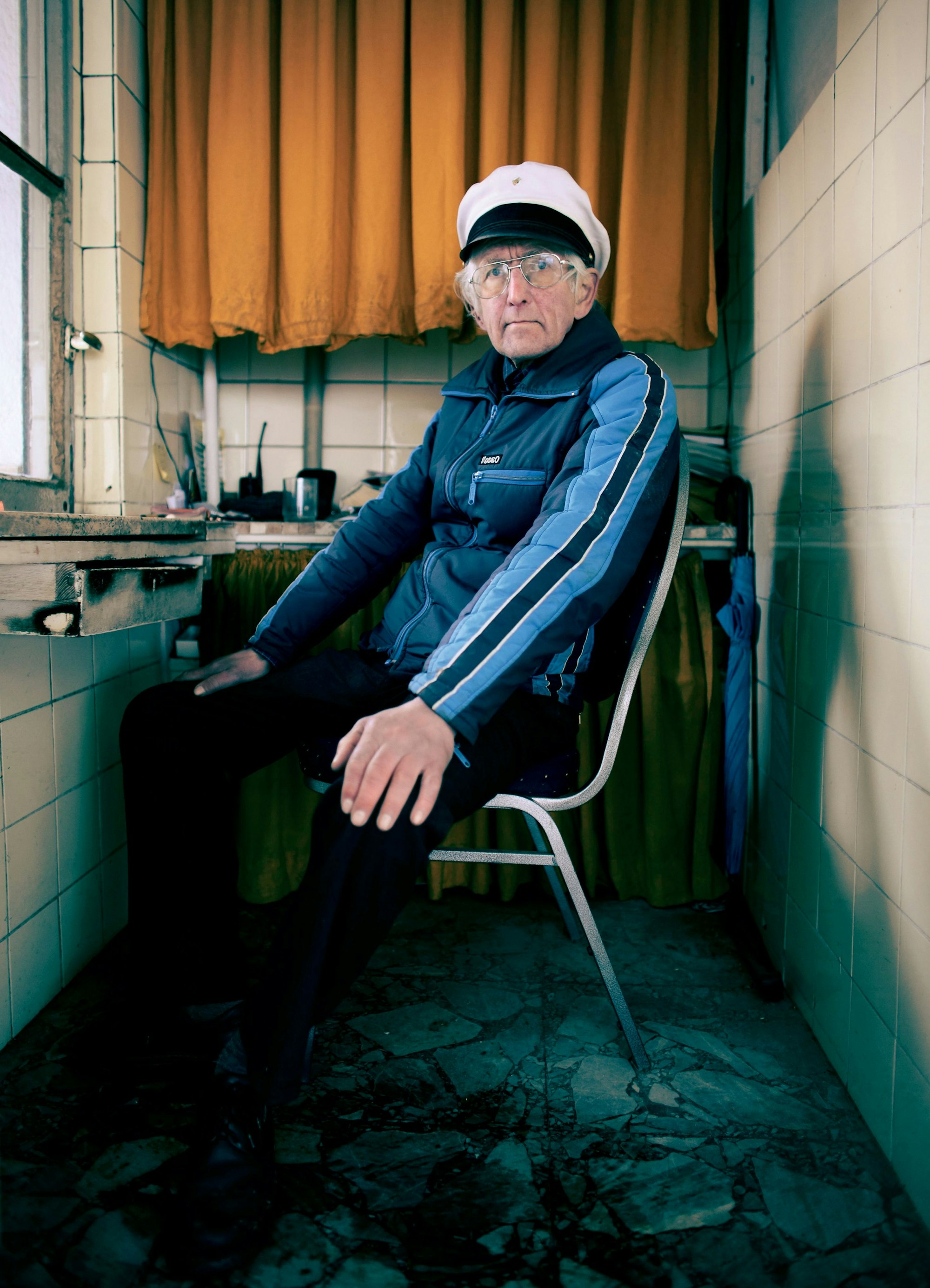 Manfred Weber sitzt mit Kapitänsmütze in seinem zweieinhalb Quadratmeter großen Büdchen. Einfach Fliesen, ein orangefarbenen Vorhang und eine kleine Anrichte sind zu sehen.