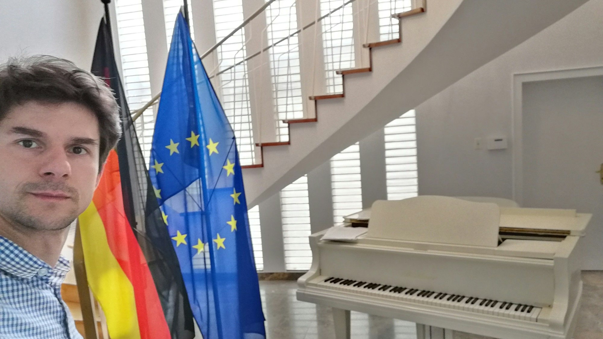 Roman Salyutov steht an einem weißen Flügel, danaben eine Deutschland- und eine Europa-Flagge.