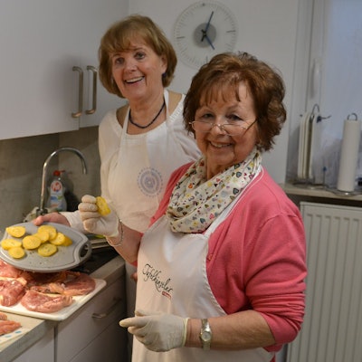 Zwei Frauen mit Schürzen stehen in einer Küche. Speisen sind auf der Arbeitsplatte und dem Schneidebrett zu sehen.