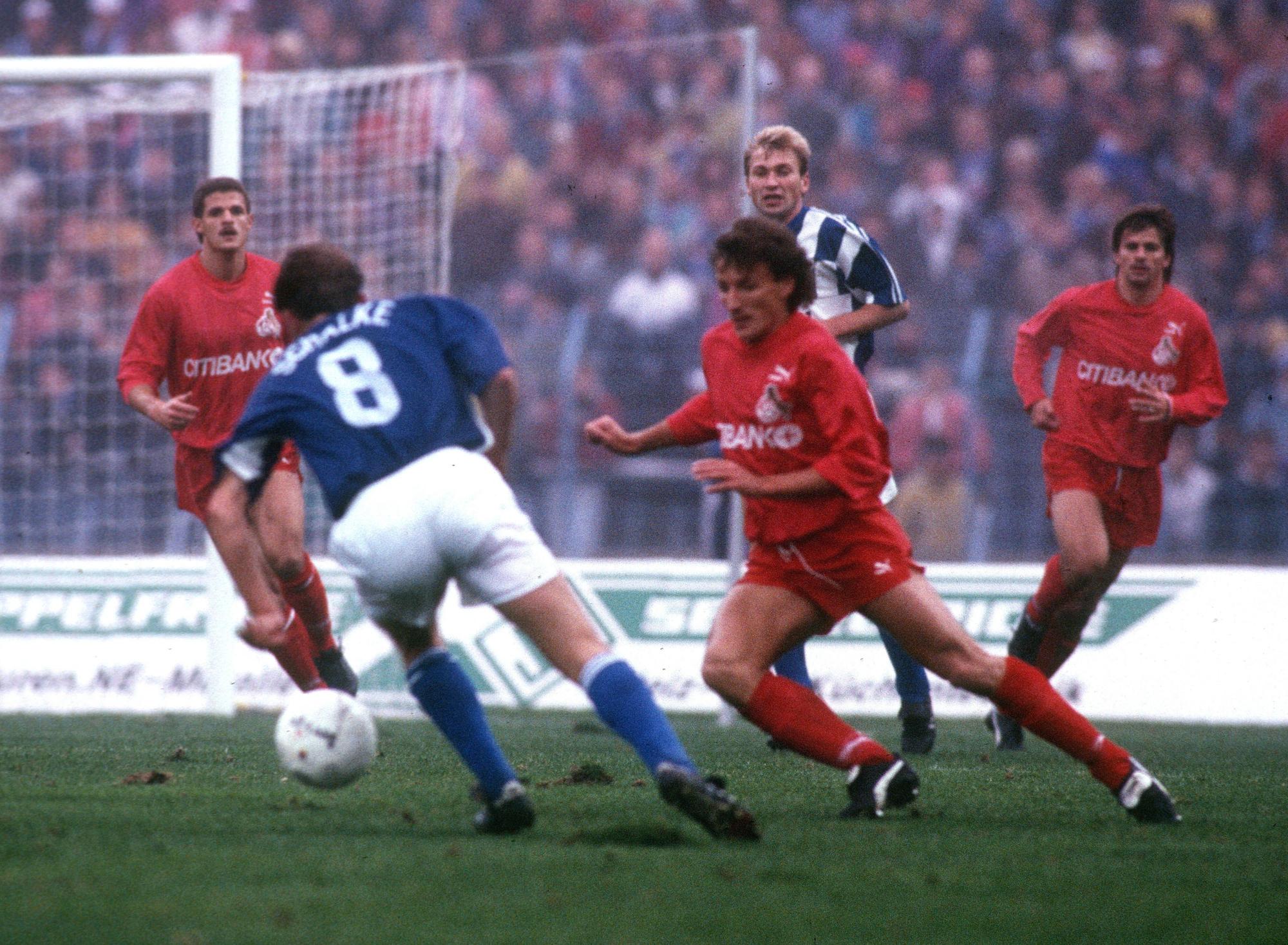 Spielszene zwischen dem FC Schalke 04 und dem 1. FC Köln. Die in Blau-Weiß gekleideten Schalker im Ballbesitz auf dem Weg zum Kölner Tor. Drei in Rot gekleidete Kölner versuchen den Ball zu erobern.