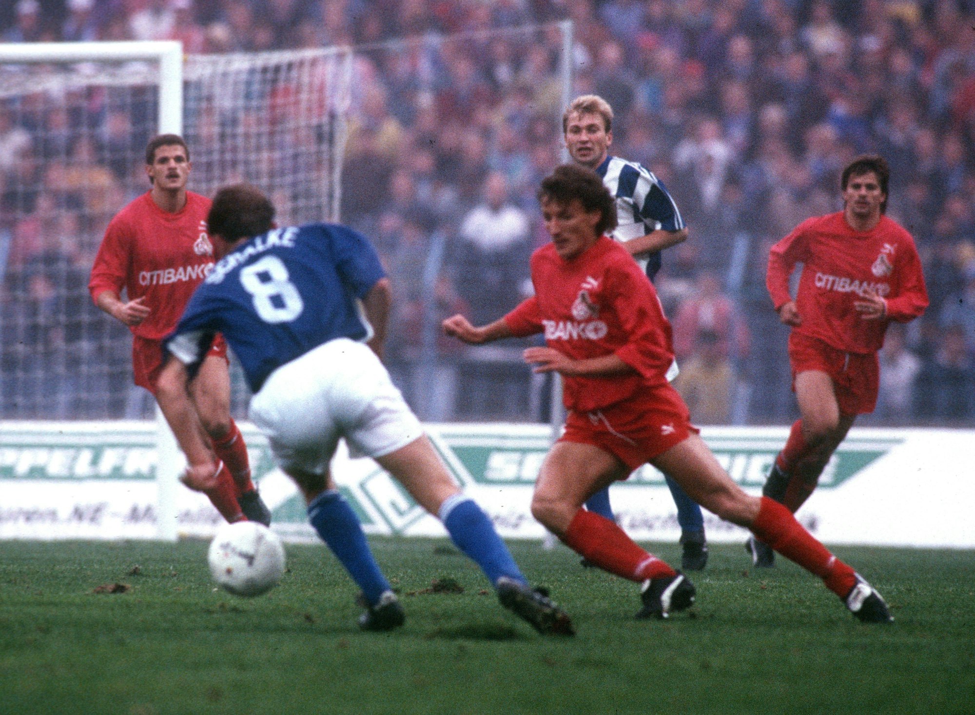 Spielszene zwischen dem FC Schalke 04 und dem 1. FC Köln. Die in Blau-Weiß gekleideten Schalker im Ballbesitz auf dem Weg zum Kölner Tor. Drei in Rot gekleidete Kölner versuchen den Ball zu erobern.