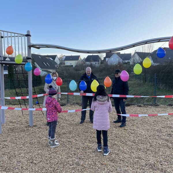 EInige Personen und Kinder stehen um ein mit Luftballons und Absperrband dekoriertes Spielplatzgerät herum.