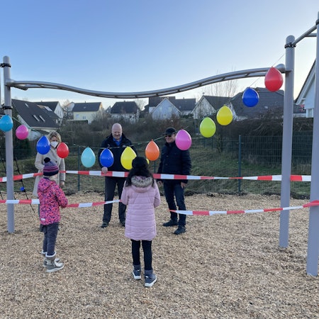 EInige Personen und Kinder stehen um ein mit Luftballons und Absperrband dekoriertes Spielplatzgerät herum.