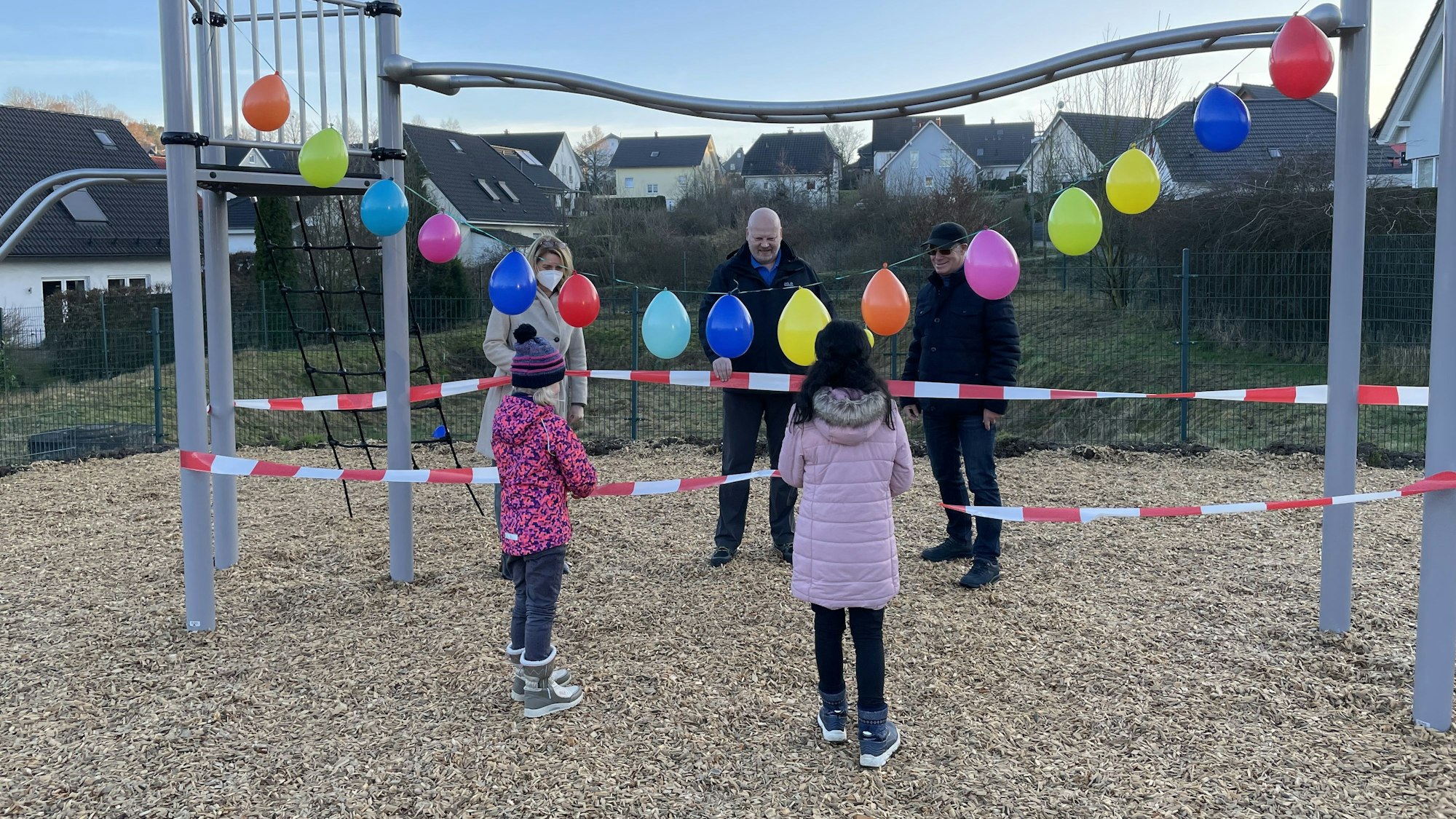 EInige Personen und Kinder stehen um ein mit Luftballons und Absperrband dekoriertes Spielplatzgerät herum.