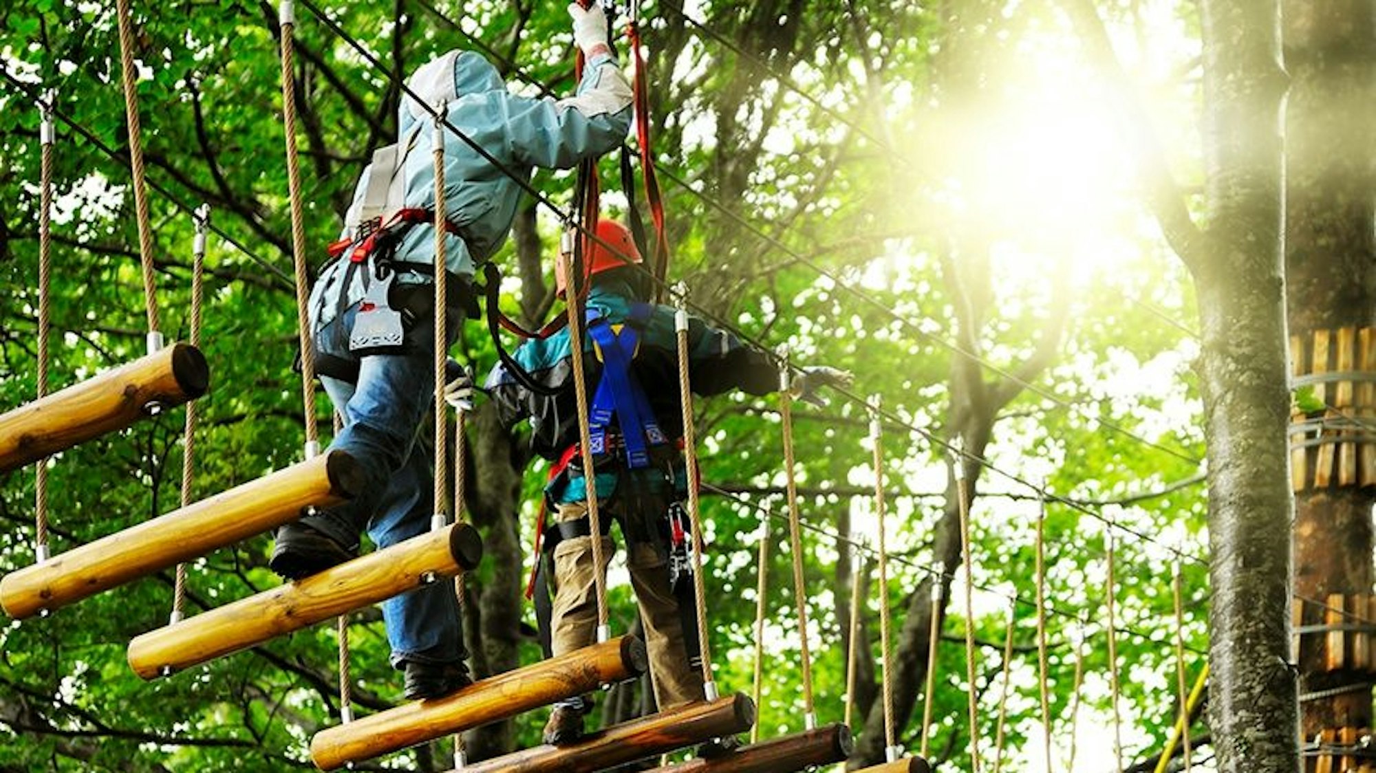 zwei Kletterer überqueren eine Route aus schwingenden Balken zwischen Bäumen