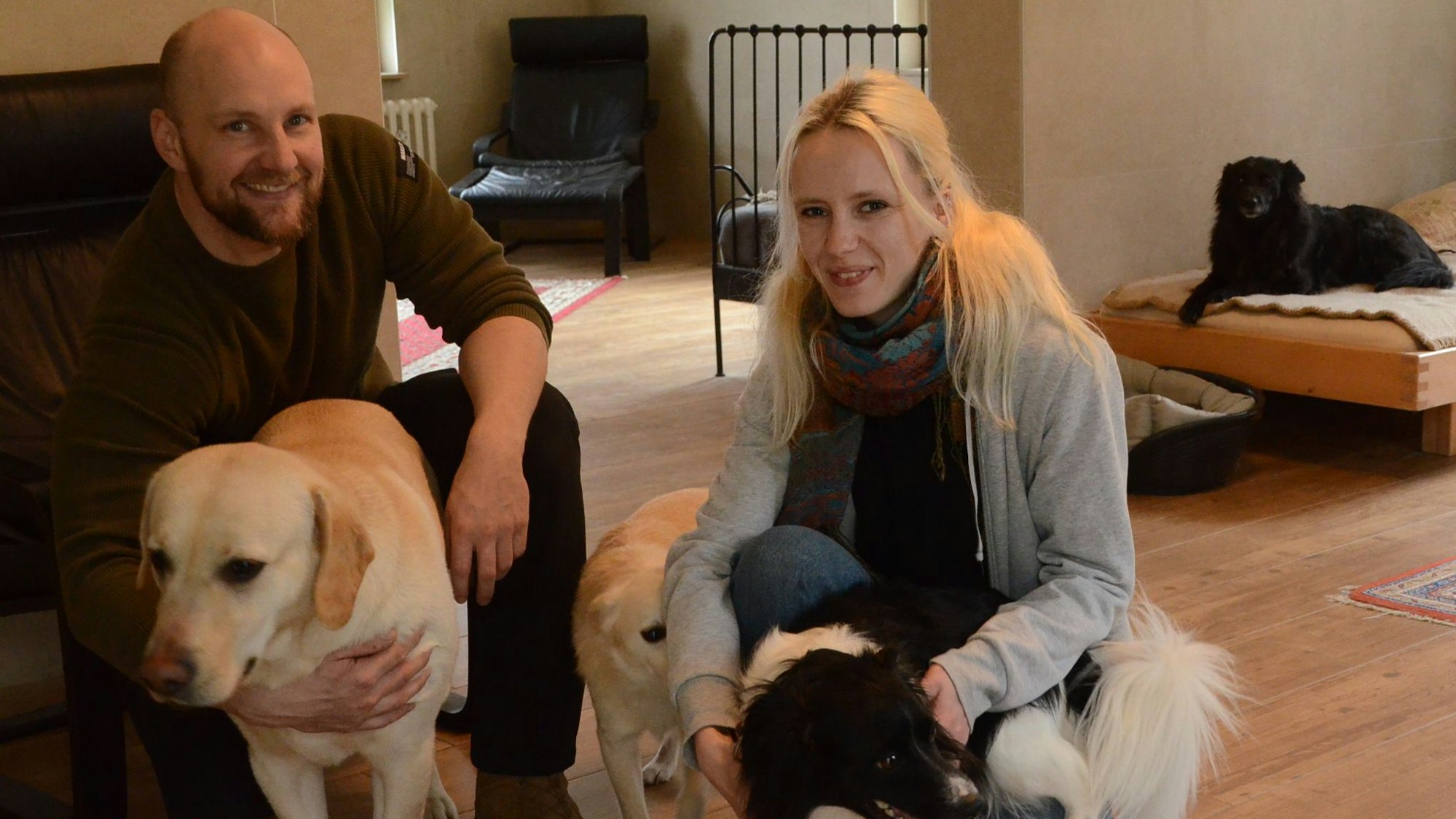 Ein Mann und eine Frau hocken am Boden; sie streicheln drei Hunde.