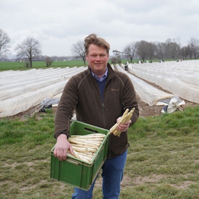 Auf dem Foto ist Johannes Nagelschmitz zu sehen. Er steht vor einem Spargelfeld und trägt eine grüne Kiste mit frisch geerntetem Spargel.