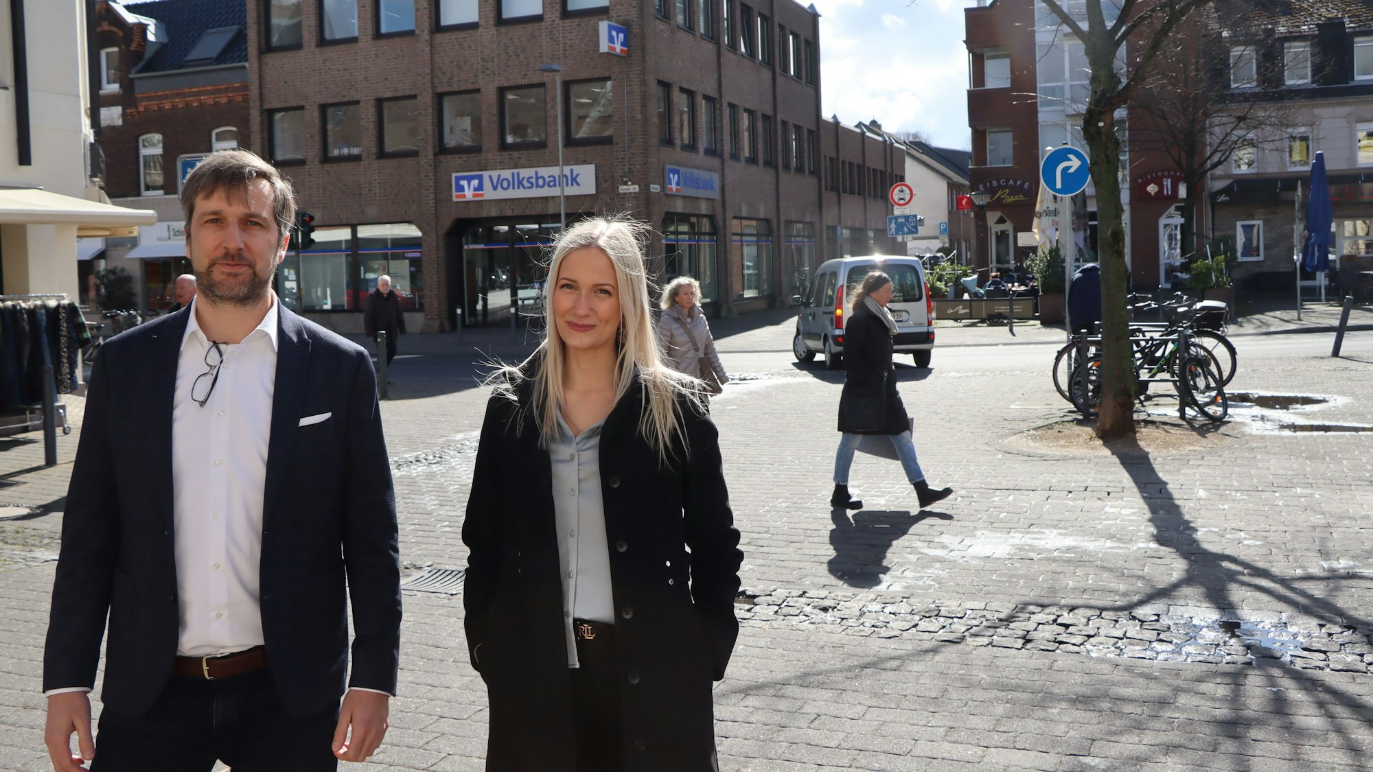 Die Juristen Alina Kriechel und Alrik Haug finden, dass das Linksabbiegeverbot schnell wieder aufgehoben werden soll. Sie stehen in Erftstadt-Lechenich. Im Hintergrund ist zu sehen, dass ein Pkw links abbiegt, obwohl das Verkehrszeichen einen Pfeil nach rechts zeigt.