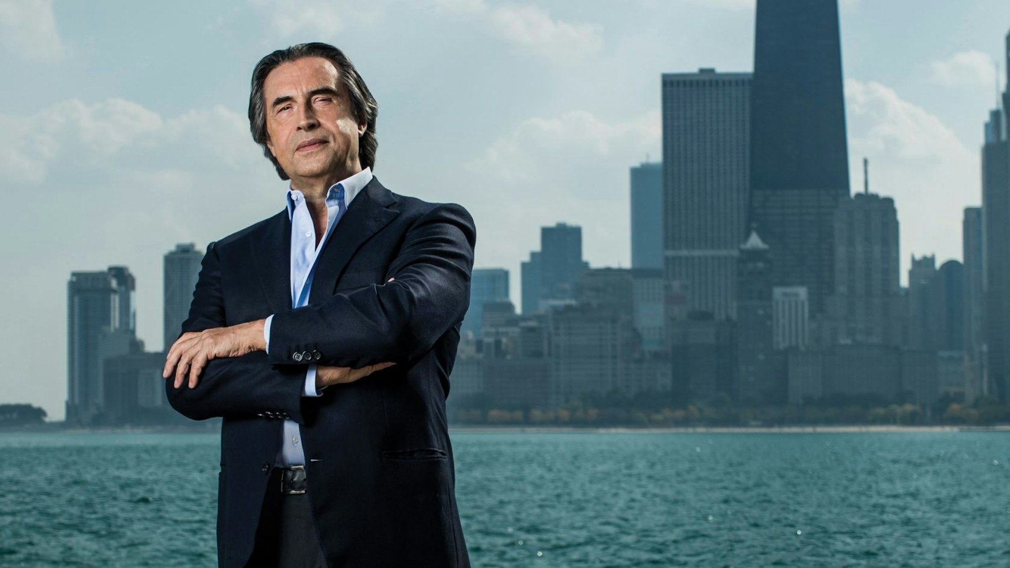 Riccardo Muti steht am Ufer des Chicago River, im Hintergrund die Stadtsilhouette.