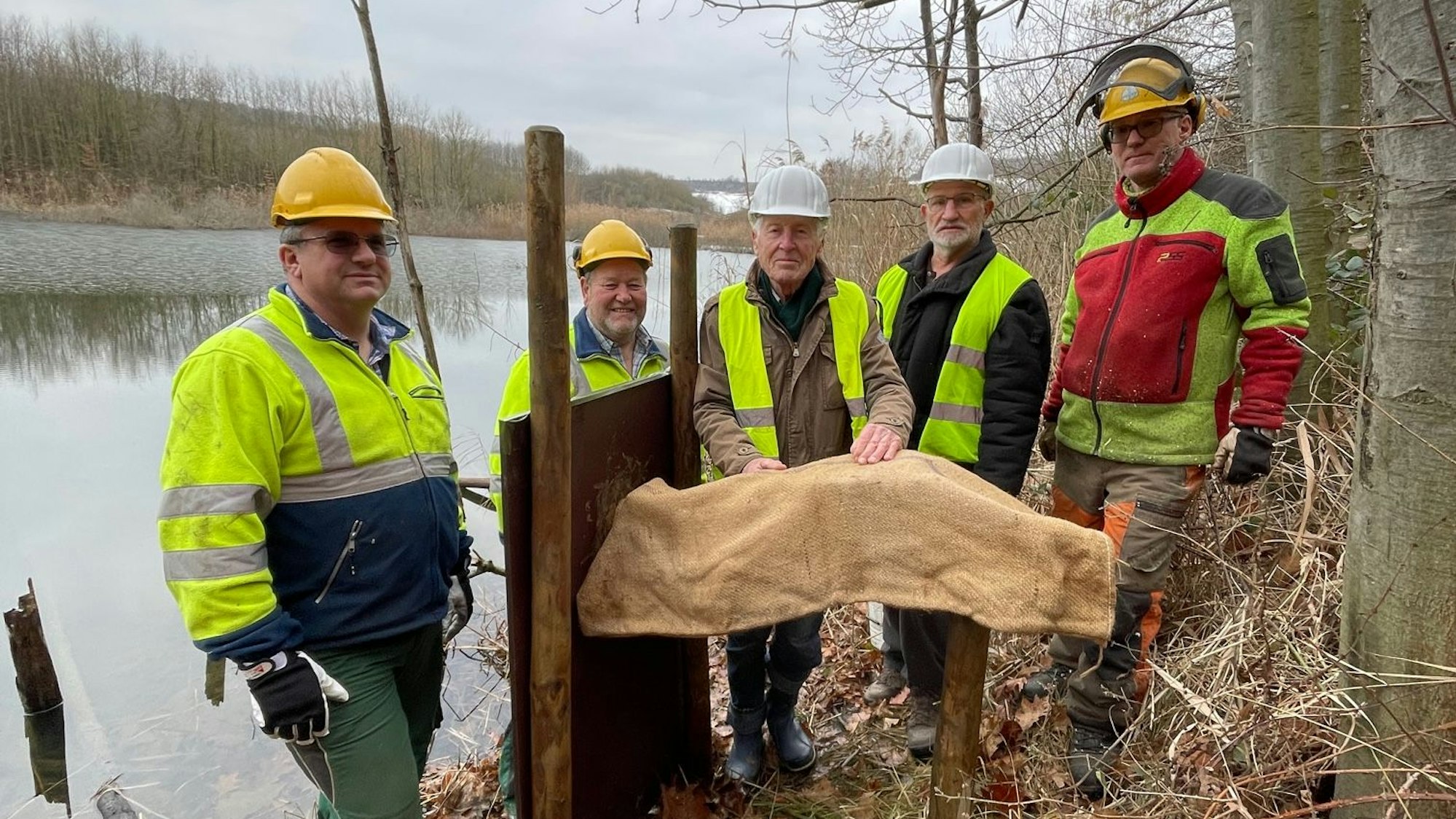 Die Männer stehen in Arbeitskleidung am Ufer eines Sees.