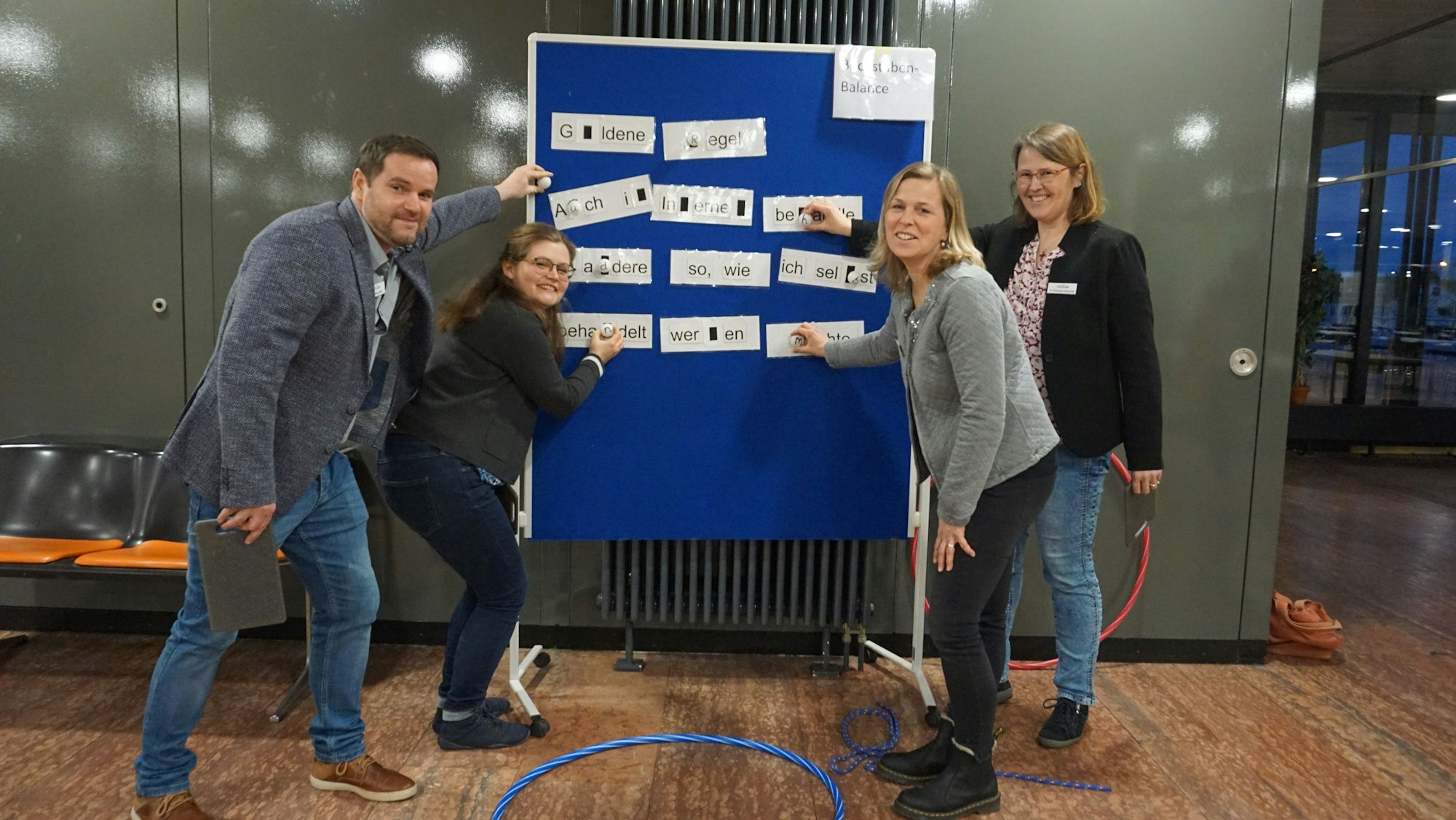 Andreas Ickes, Jana Wiens, Heike Heinen und Svenja Pommer-Schmitt stehen vor einer blauen Tafel, welche das Konzept für den "Medienparcours" für dritte und vierte Klassen präsentiert.