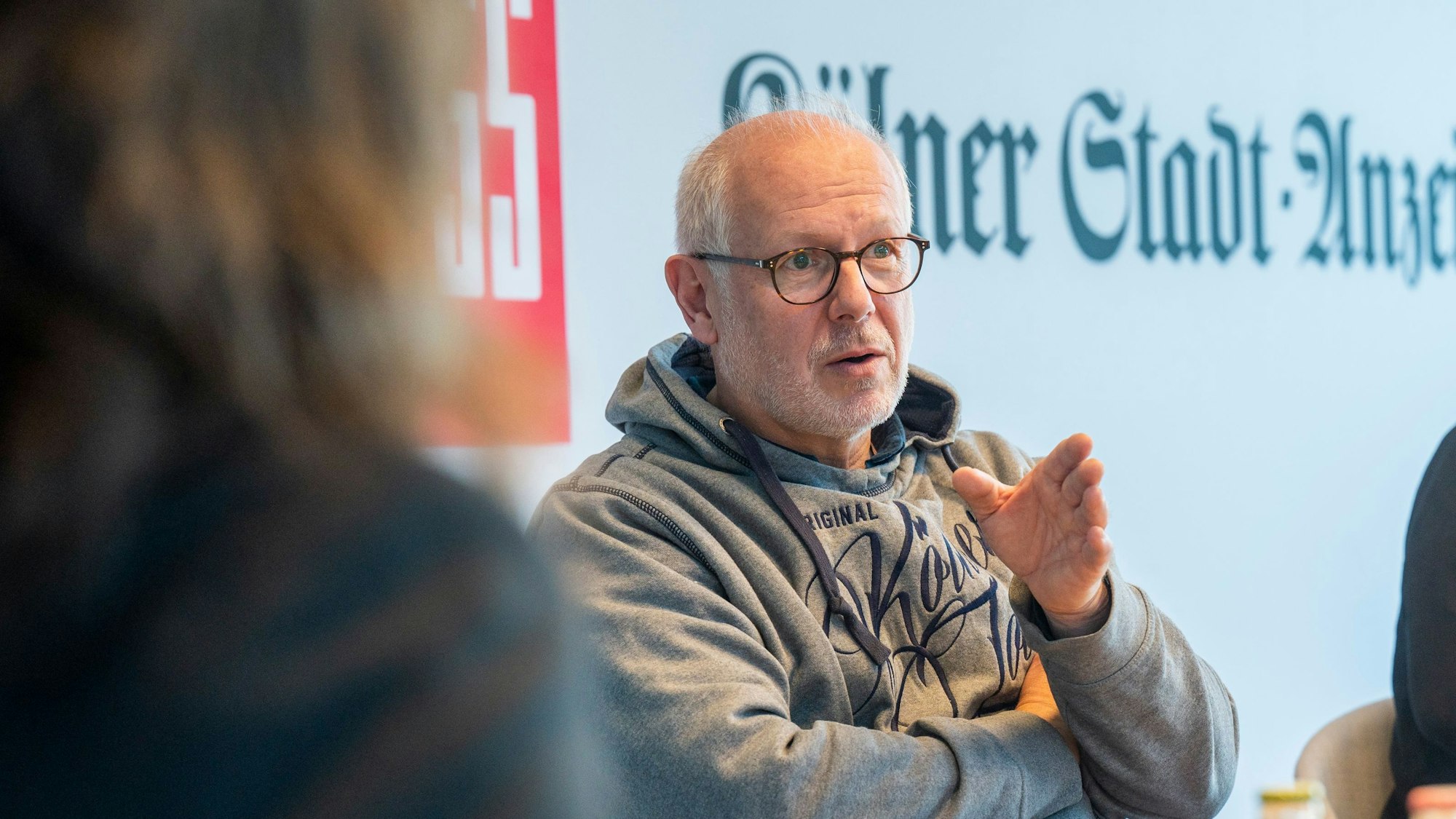 Theo Pagel spricht und gestikuliert mit der linken Hand. Er hat weißes Haar und einen kurzen weißen Bart, er trägt eine Brille und einen grauen Kapuzenpullover.