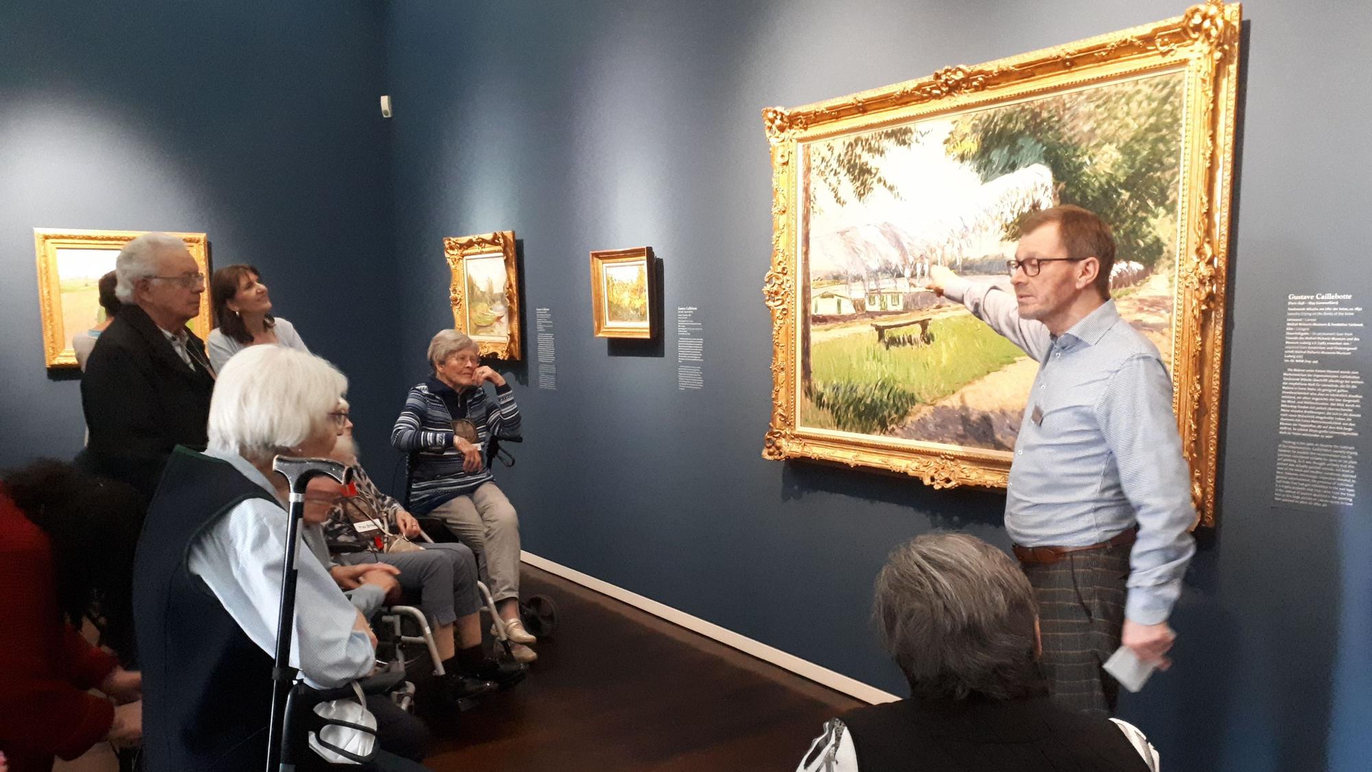 Ein Mann beschreibt ein Kunstwerk in einem Museum. Seniorinnen und Senioren schauen ihm zu.