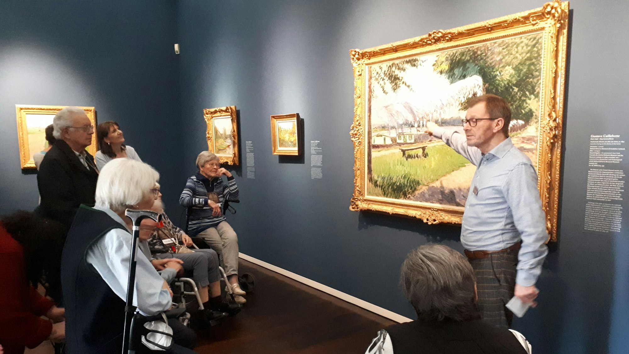 Ein Mann beschreibt ein Kunstwerk in einem Museum. Seniorinnen und Senioren schauen ihm zu.