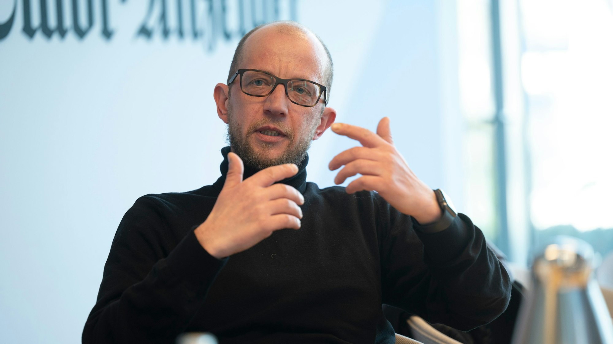 Simon Darscheid ist in einer Gesprächssituation zu sehen, er gestikuliert und schaut sein Gegenüber an. Er trägt eine Brille und einen schwarzen Rollkragenpullover.