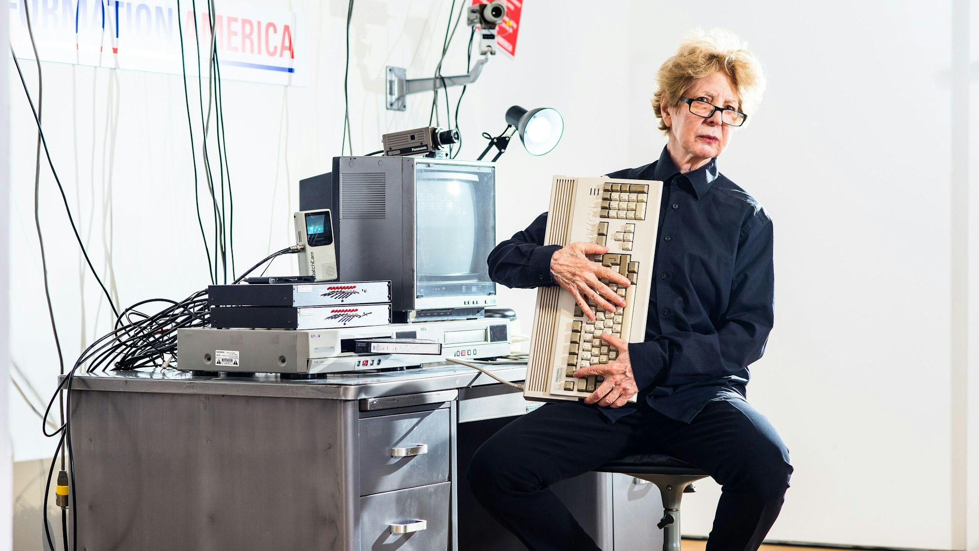Julia Scher sitzt in ihrer Installation Information America und hält eine alte Computertastatur in den Händen.