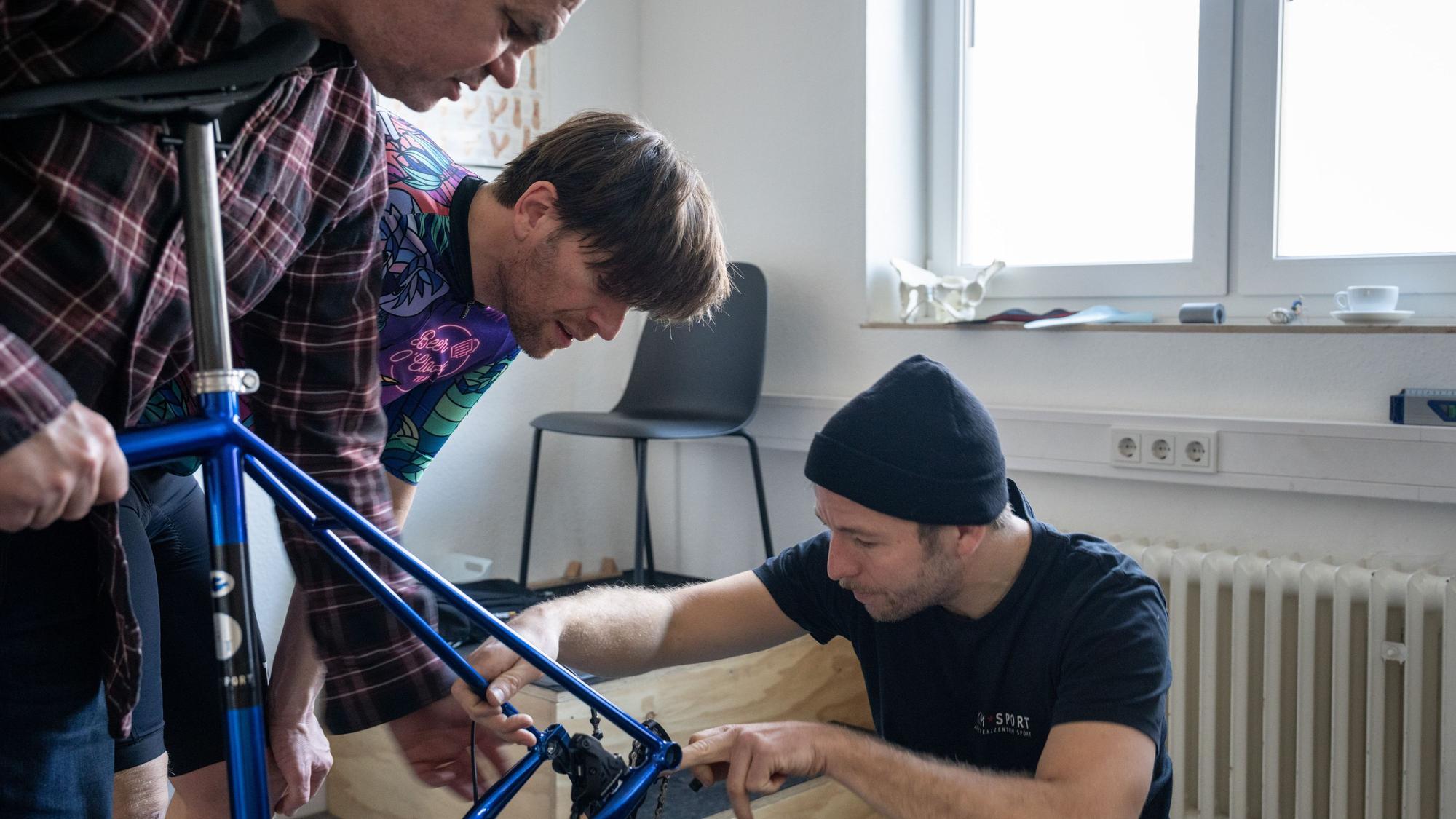 James Buckley, Tim Farin und Sebastian Klaus beugen sich über den Rahmen eines Fahrrads.