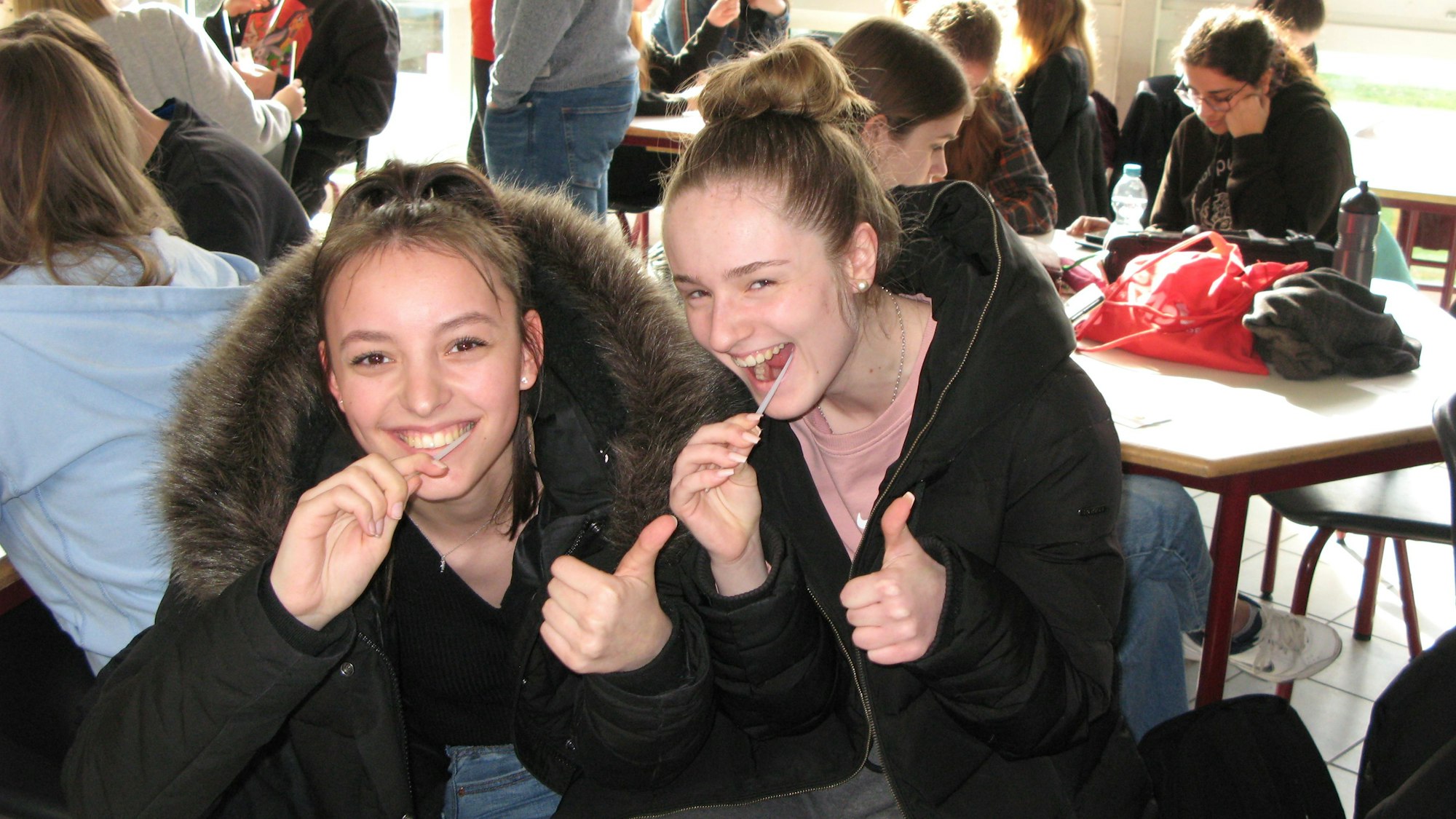 Zwei Mädchen sitzen an einem Tisch, eine hat ein Wattestäbchen im Mund.