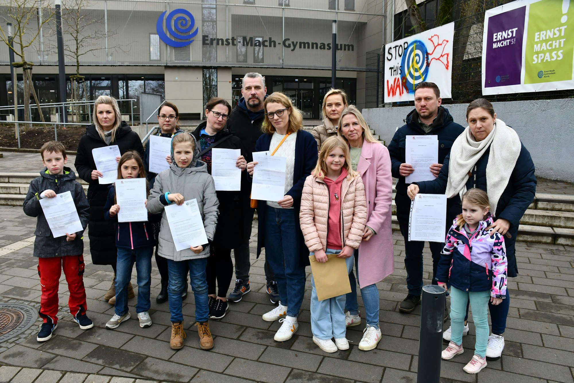 Eine Gruppe von Eltern und Kindern halten Ablehnungsschreiben in den Händen. SIe stehen vor dem Eingang des Ernst-Mach-Gymnasiums.