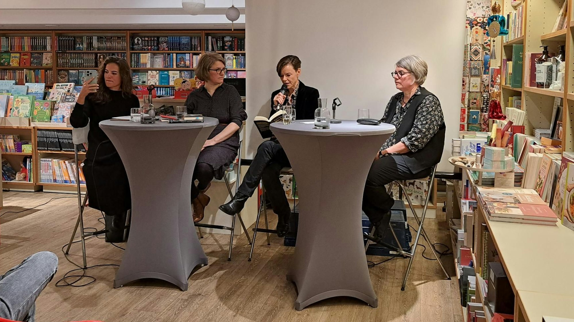 Moderatorin Mona Ameziane (links), Johanna Schwering, Antje Rávik Strubel und Birgit Weyhe sitzen an zwei Hochtischen im Buchladen Neusser Strasse. Ameziane nimmt ein Video auf. Rávik Strubel liest aus dem Buch „Wer hat Bambi getötet?“, das sie übersetzt hat. Schwering und Weyhe hören zu.