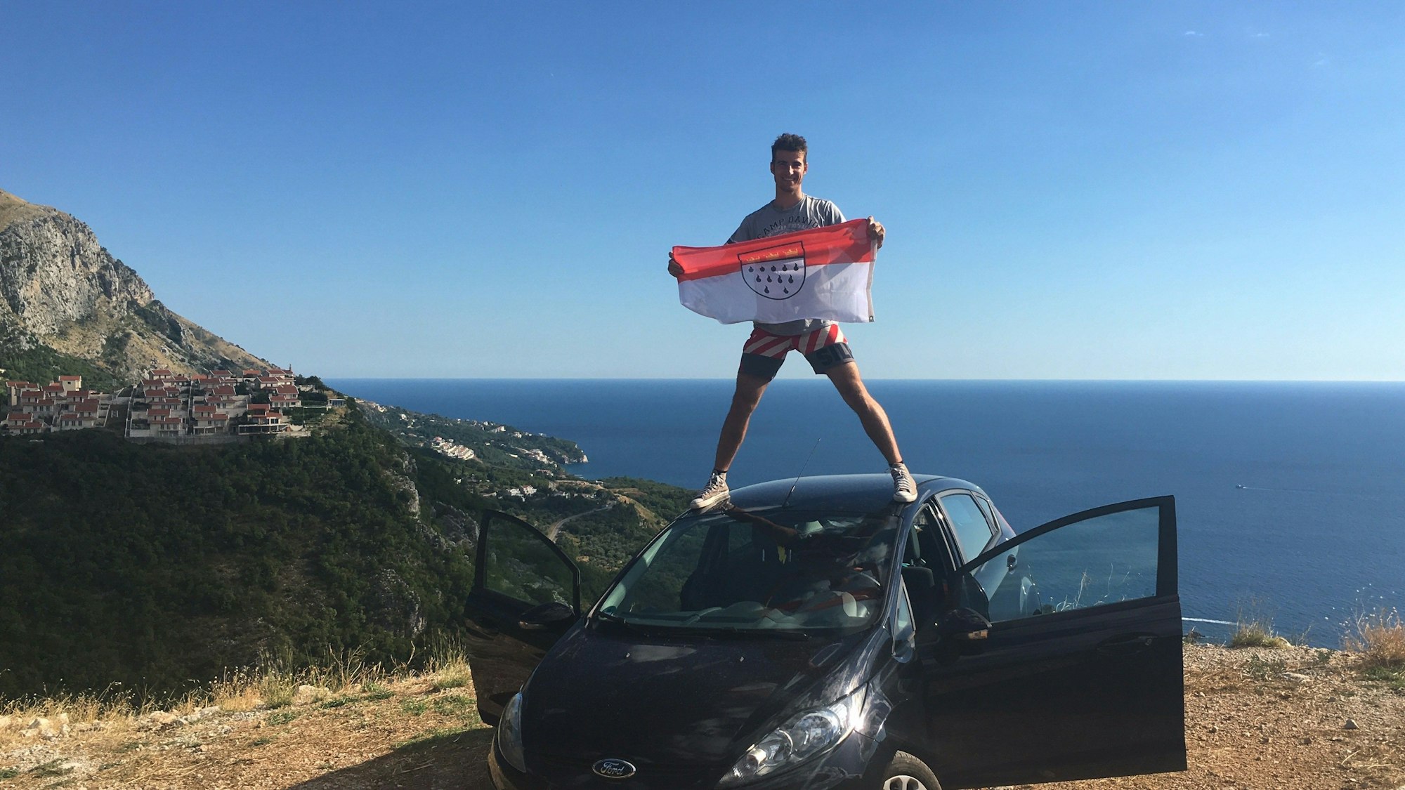 Niklas Bahn steht in Montenegro auf dem Dach seines schwarzen Autos und hält die Köln-Flagge in den Händen. Hinter ihm ist das Meer.