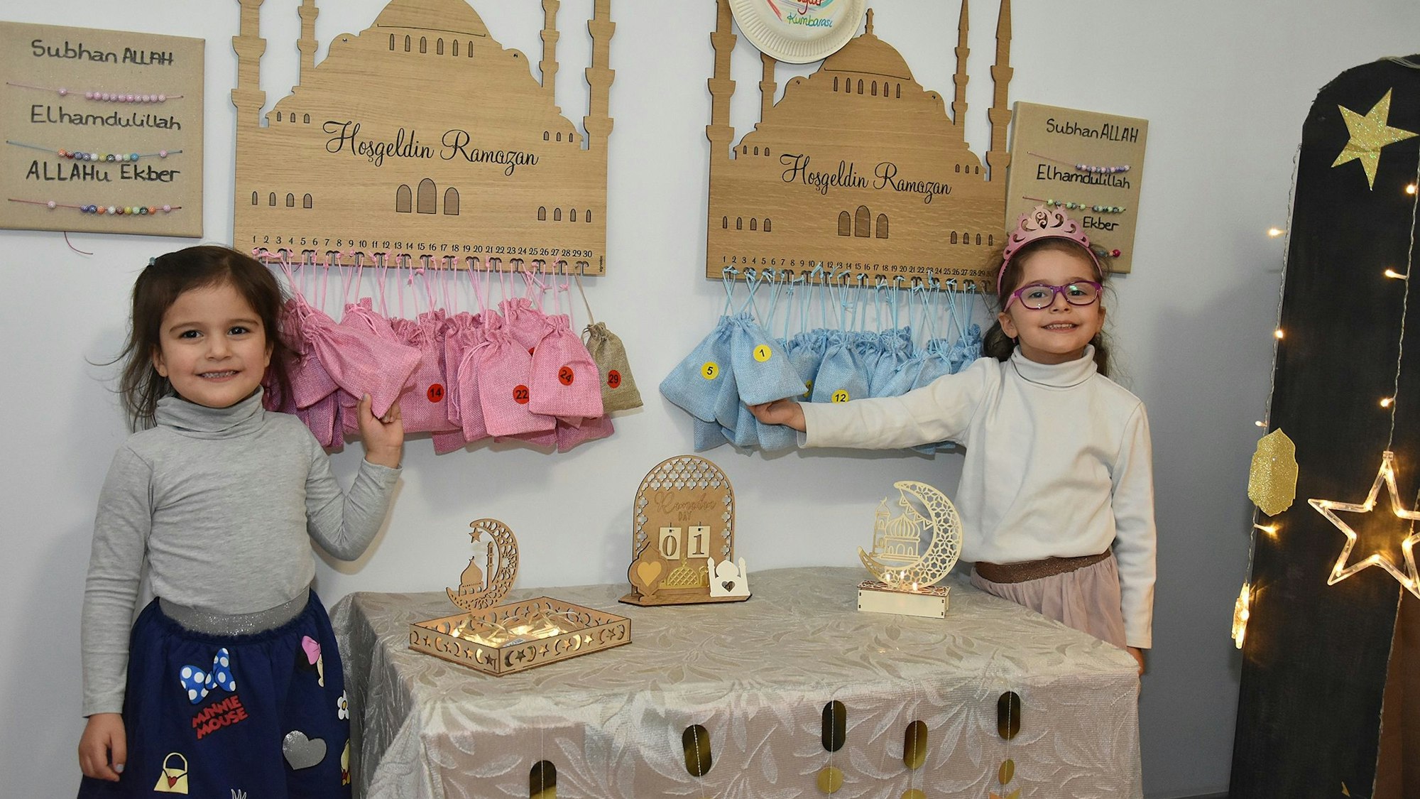 Zwei kleine Mädchen stehen vor Ramadan-Dekorationen. In dreißig Säckcken, die unter einem hölzernen Moschee-Schild aufgehangen sind, erwarten sie Geschenke.