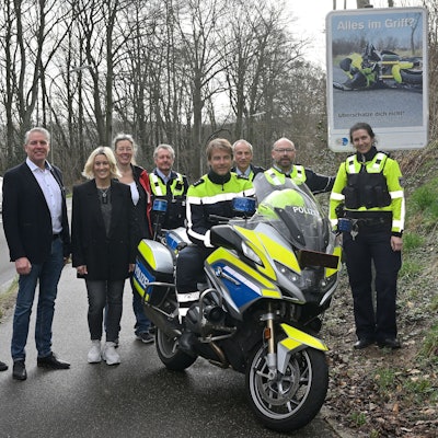 Polizeibeamtinnen und -beamte sowie Vertreter von Gemeinden, Städten und Kreis stehen vor einem neuen Schild, das Motorradfahrer zu Vorsicht anhalten soll. Über dem Bild eines verunglückten Motorrads ist der Schriftzug„ Alles im Griff?“ zu lesen.