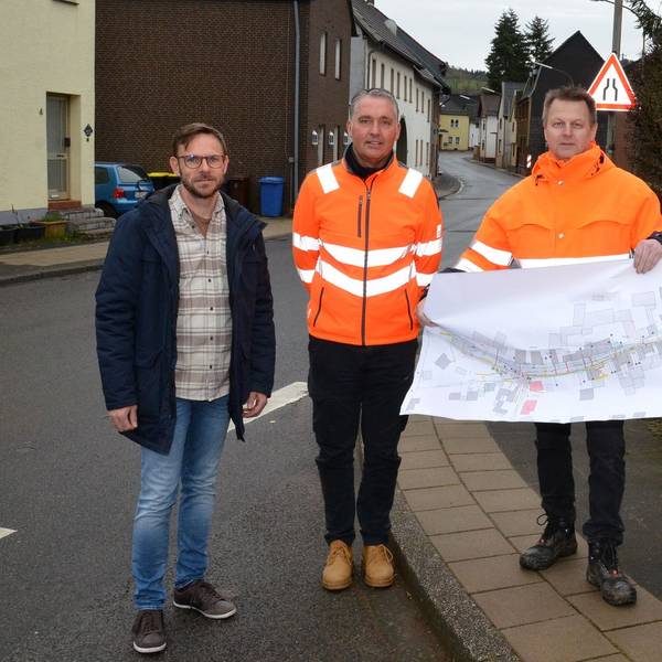 Drei Männer mit orangen Warnwesten halten einen Stadtplan hoch. Daneben steht ein Mann in Straßenklamotten.