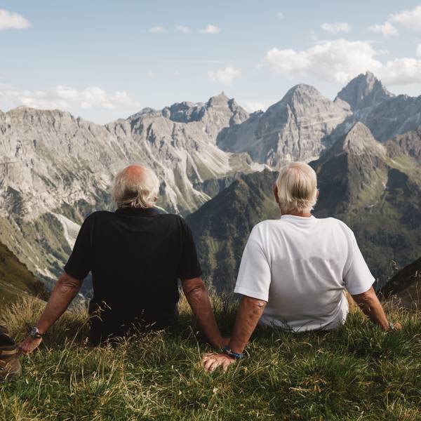 Zwei ältere Menschen sitzen an einem Hang und schauen auf eine Berglandschaft hinaus.