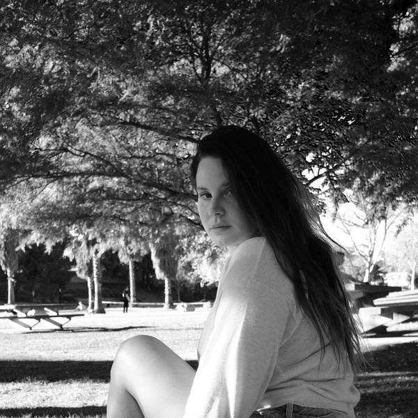 Lana Del Rey sitzt in einem Park unter einem Baum und schaut in die Kamera. Das Bild ist schwarz-weiß.