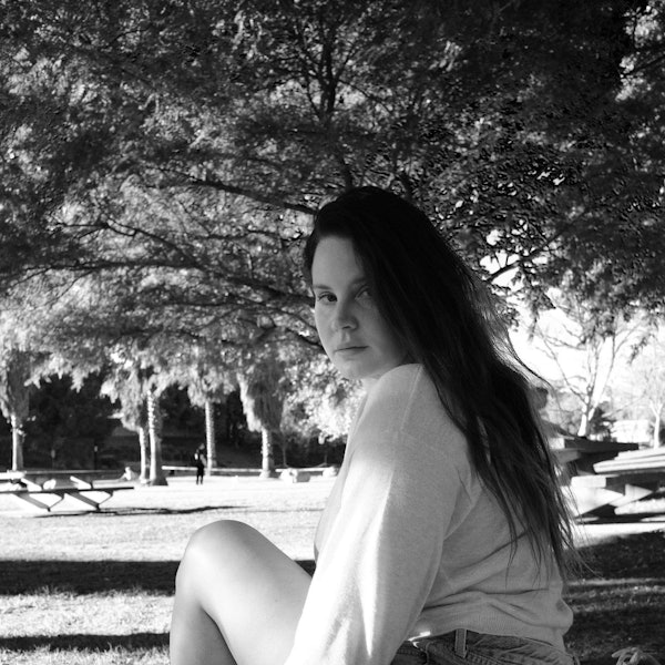 Lana Del Rey sitzt in einem Park unter einem Baum und schaut in die Kamera. Das Bild ist schwarz-weiß.