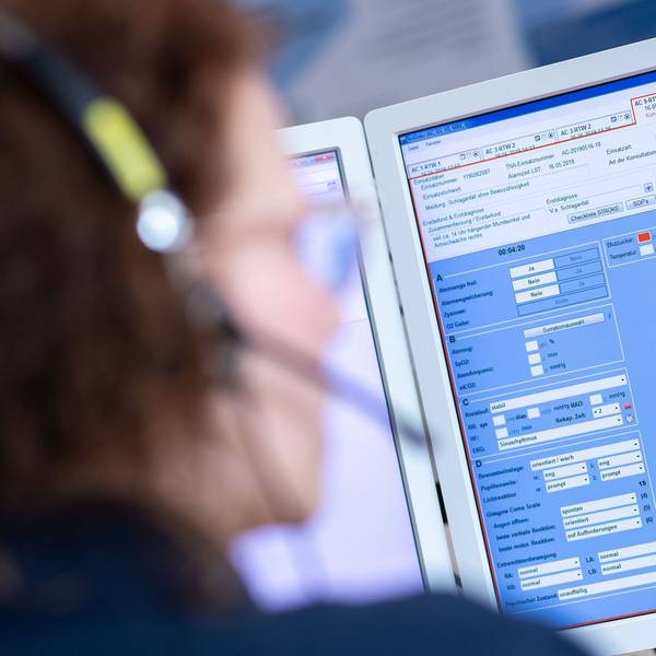 Eine Telenotärztin sitzt in der Rettungsleitstelle vor einem Bildschirm und ist mit Rettungssanitätern verbunden, die eine Schlaganfallpatientin behandeln.